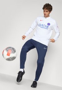 Bijela dugacima rukava sportska majica s logotipom "QATAR AIRWAYS" i plavim detaljima, uparen s mornarskoplavim trenirkama i crnim loptačkim cipelama, uz nogometnu loptu.