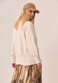 Suéter acolchado de color crema con mangas texturizadas, combinado con una falda fluida y estampada en tonos terrenales y una gorra de béisbol color tostado.