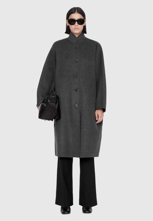 Personne portant un manteau gris charbon boutonné, un pantalon noir à jambes larges, des bottes noires, de grandes lunettes de soleil noires, et tenant un sac à main noir.