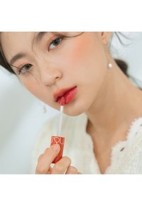 Lucidalabbra rosso in un tubo trasparente con applicatore a pennello. Soggetto che applica il lucidalabbra sulle labbra. Maglione leggero, sfondo morbido. Luce naturale utilizzata.