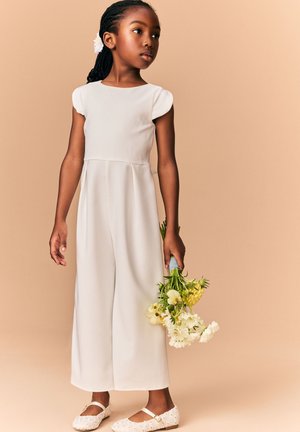 Giovane ragazza in abito bianco che tiene un mazzo di fiori gialli e bianchi su sfondo beige.