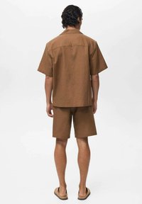 Cămașă scurtă maro cu mânecă scurtă, asociată cu pantaloni scurți asortați, realizată dintr-un material texturat. Afișată din spate, arătând o croială lejeră.