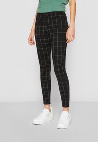 Sorte stretchy leggings med et hvitt rutenettmønster, med elastisk midjebånd og slim fit, paret med hvite sneakers.
