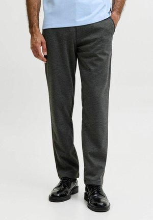 Mann trägt dunkelgraue Hose und glänzende schwarze Anzugschuhe und steht mit einer Hand in der Tasche vor schlichtem Hintergrund.