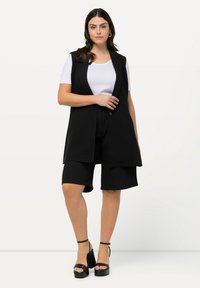 Zwarte mouwloze blazer over een witte top, gecombineerd met zwarte op maat gemaakte shorts. Het model draagt zwarte platform sandalen. Glad materiaal, gestructureerd ontwerp.