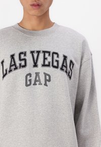 Grå genser laget av en bomullsblanding med "LAS VEGAS" og "GAP" trykt i fet svart og grå skrift over fronten.