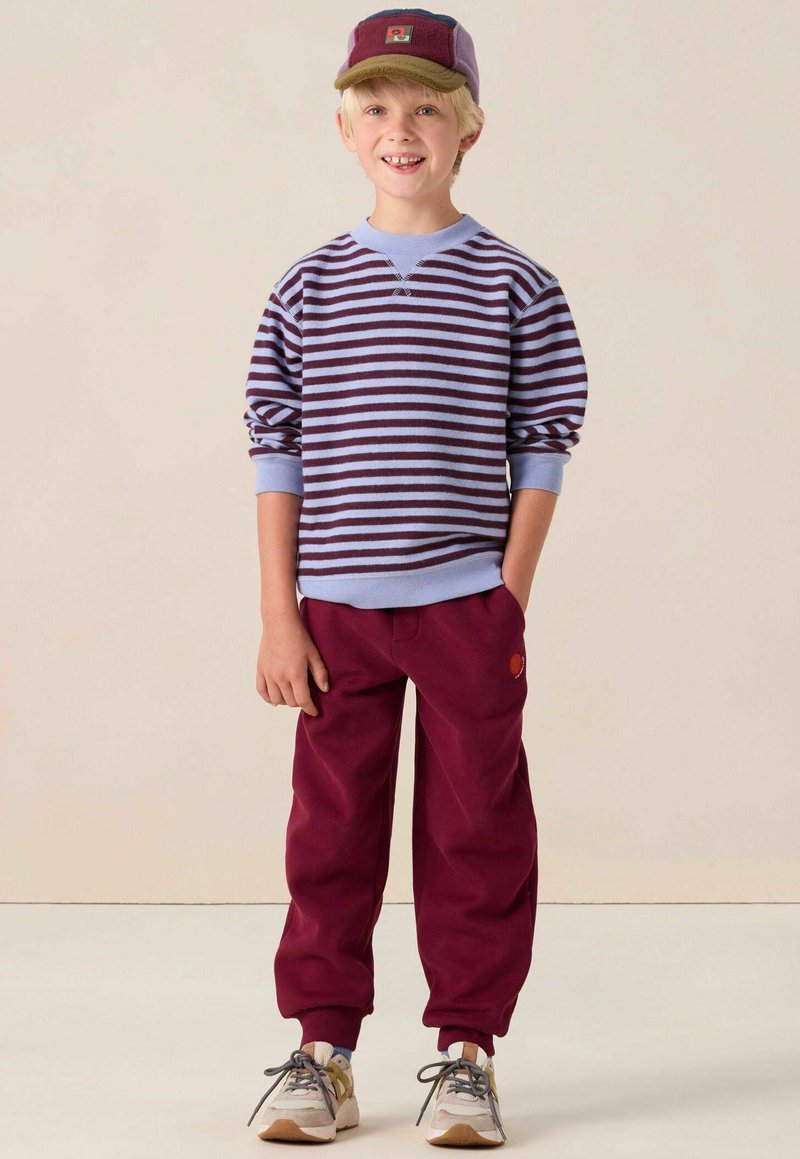 Pull rayé en lavande et violet foncé, associé à un pantalon bordeaux et des baskets beiges. La casquette présente un design multicolore avec un patch.