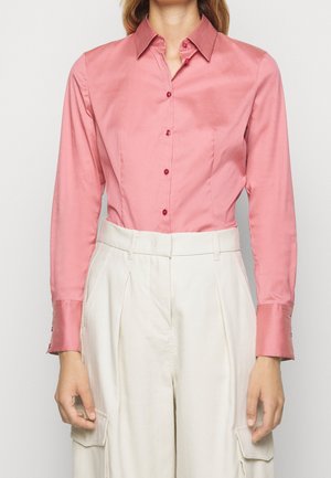 Femme portant une chemise rose ajustée boutonnée à manches longues et un pantalon blanc taille haute avec poches cargo, debout devant un fond uni.