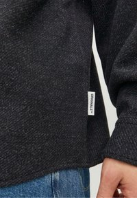 Felpa nera con texture e orlo curvo, con etichetta visibile etichettata "ORIGINALS". Il tessuto ha un aspetto morbido e intrecciato.