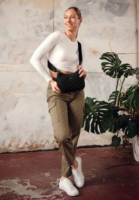 Černá prošívaná kabelka přes rameno s zakřiveným tvarem, nastavitelným popruhem a embosovaným logem, spárovaná s olivově zelenými cargo kalhotami a bílými teniskami.