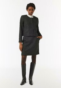 Czarna marynarka damska z tkaniny tweed, z okrągłym dekoltem i dwoma kieszeniami, w zestawieniu z dopasowaną spódnicą ołówkową. Całość dopełniają czarne, skórzane kozaki za kolano.