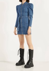 Vestido de mezclilla con mangas largas, cremallera frontal y cintura ceñida con cinturón. Presenta un cuello con solapa y un diseño de hombros abullonados. Se lleva con botas de combate negras con cordones.