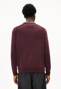 Mann mit kurzen, lockigen Haaren, der einen einfarbigen, kastanienbraunen Pullover und dunkle Jeans trägt, steht mit dem Rücken zu einem weißen Hintergrund.
