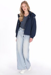 Granatowa wodoodporna kurtka z kapturem, noszona na koszuli w paski, w zestawieniu z jasnoniebieskimi szerokimi dżinsami i białymi trampkami.