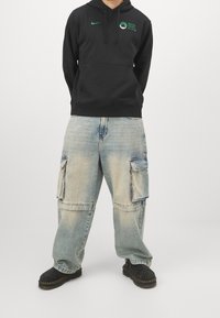 Sweat à capuche noir Nike avec logo vert, associé à un jean cargo ample bleu clair avec poches latérales et poignets élastiques, porté avec des bottes noires.