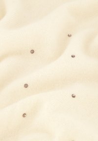 Cremfarbener gestrickter Stoff mit einer weichen Textur und kleinen metallischen Nieten, die gleichmäßig über die Oberfläche verteilt sind.