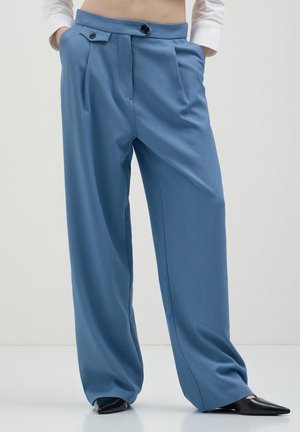 Helleblaue weite Hosen mit Falten, die zwei Vordertaschen und einen Knopfverschluss aufweisen. Glatte Textur und taillierte Passform. Schwarze spitze Schuhe.