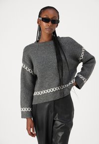 Grau gestrickter Pullover mit weißen Kreuzstichdetails, kastiger Form und weiten Ärmeln, kombiniert mit schwarzen Lederhosen.
