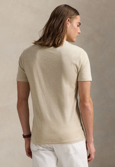 Mladý muž so stredne dlhými hnedými vlasmi, oblečený v béžovom tričku s krátkym rukávom a bielych nohaviciach, zobrazený zozadu na neutrálnom pozadí.