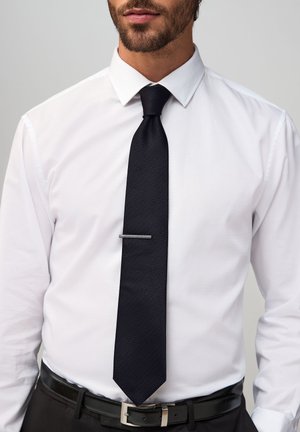 Man wearing white dress shirt, black textured tie with tie clip, and black leather belt against a plain gray background.