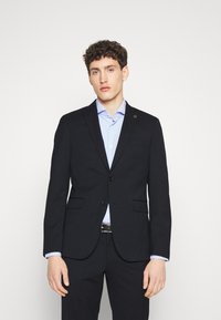 Blazer blu scuro con taglio sartoriale, due bottoni, revers a punta e tasche aperte, abbinato a una camicia a quadri azzurri.