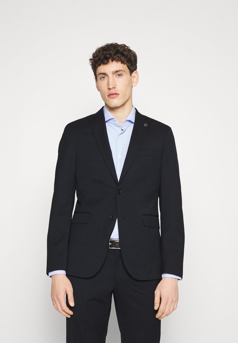 Blazer blu scuro con taglio sartoriale, due bottoni, revers a punta e tasche aperte, abbinato a una camicia a quadri azzurri.