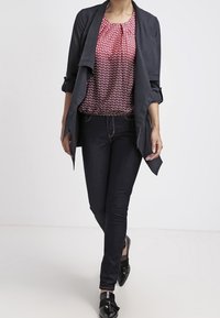 Giacca nera drappeggiata sopra una blusa rossa e bianca a fantasia, abbinata a jeans skinny scuri e mocassini neri. Tessuto morbido e design casual.