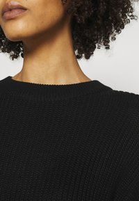 Pull noir en maille avec un col rond, présentant un motif texturé et des poignets côtelés, positionné près de la clavicule.