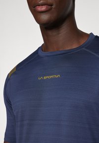 Tumši zils sporta krekls ar īsām piedurknēm, teksturēts audums un dzeltenais "LA SPORTIVA" logo uz krūšu. Ergonomisks dizains.