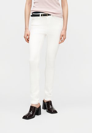 Persona che indossa pantaloni bianchi aderenti, top rosa chiaro infilato dentro, cintura nera e mules con tacco marrone scuro, in piedi su uno sfondo semplice.