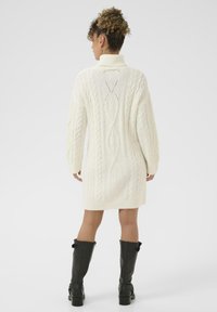 Abito maglione cream a coste con colletto alto, caratterizzato da un design a motivi sulla schiena. Indossato con stivali neri sopra il ginocchio per un contrasto.