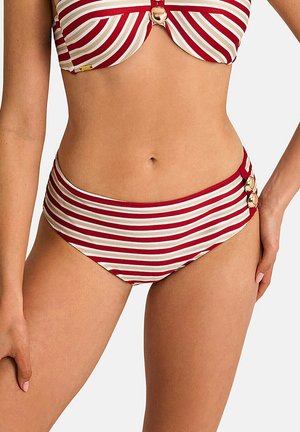 Femme portant un bikini rayé rouge et blanc avec des détails de boutons dorés sur le haut et le bas, debout devant un fond uni.