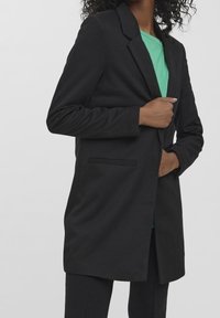 Blazer nero realizzato in tessuto liscio con chiusura a un bottone, dotato di revers classici e tasche laterali. Indossato sopra una camicia verde.
