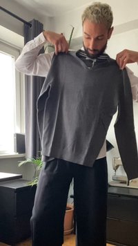 Gray knitted sweater held up, featuring a V-neck and long sleeves. The model wears a white shirt underneath and dark trousers.