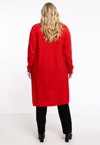 Cardigan long rouge à manches longues, avec une texture lisse et des fentes latérales, associé à un pantalon noir et des bottes à talons noires.