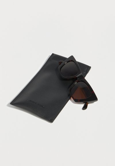 Μαύρα γυαλιά ηλίου σε σχήμα γάτας με σκουρόχρωμους φακούς, ακουμπισμένα σε μαύρη δερμάτινη θήκη πάνω σε λευκή επιφάνεια.