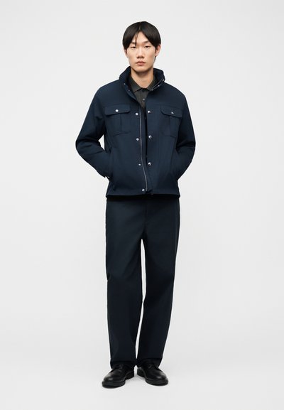 Νεαρός άνδρας όρθιος με τα χέρια στις τσέπες, φοράει σκούρο μπλε μπουφάν, μαύρο παντελόνι, μαύρο πουκάμισο και μαύρα παπούτσια, μπροστά σε απλό φόντο.