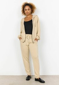 Hoodie beige e pantaloni jogger abbinati realizzati in materiale morbido, con vita regolabile, dettagli con zip e polsini a coste. Top e scarpe neri.