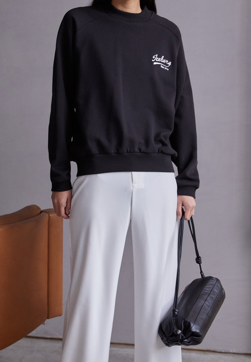 Sweat-shirt noir à col rond et manches raglan, avec un petit logo blanc. Associé à un pantalon blanc à jambes larges et un sac noir.