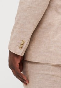 Manga de chaqueta de traje de lino beige con tres botones marrón claro, que muestra una tela texturada y un corte a medida. Mano posicionada cerca del puño.