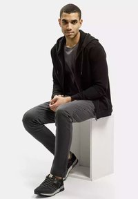 Czarna bluza z kapturem na suwak na szarym t-shircie, szare spodnie slim fit i czarne trampki z akcentami, siedząca na białym sześcianie.