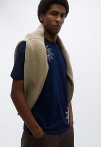 Μπλε σκούρο μπλουζάκι με κέντημα ήλιου, σε συνδυασμό με ανοιχτό μπεζ πουλόβερ πλεκτό που είναι περασμένο πάνω από τους ώμους. Καφέ παντελόνι συμπληρώνει την εμφάνιση.