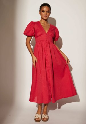 Abito lungo in rosso vivace, tessuto strutturato con maniche a sbuffo, scollatura a V profonda e chiusura frontale con bottoni. Sandali bianchi completano il look.