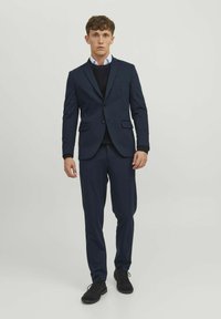 Giacca su misura blu navy con taglio slim, dotata di blazer a un bottone e pantaloni abbinati, abbinata a un maglione nero lavorato a maglia e scarpe nere.