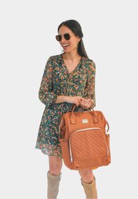Mochila de cuero acolchado naranja con asas dobles, bolsillo frontal con cremallera y bolsillos laterales. El modelo lleva puesto un vestido floral y gafas de sol.