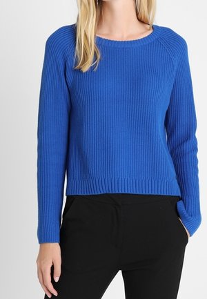 Femme portant un pull bleu côtelé à manches longues avec un col rond et un pantalon noir, debout sur un fond blanc uni.