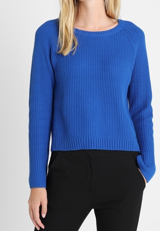 Femme portant un pull bleu côtelé à manches longues avec un col rond et un pantalon noir, debout sur un fond blanc uni.