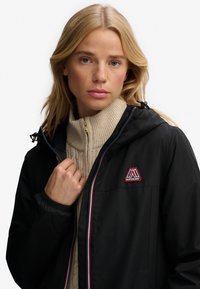 Sort vandtæt jakke med hætte, der har en lynlåslukning og et logo med røde accenter. Båret over en lys beige striktrøje.