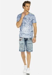 Hellblaues kurzärmliges T-Shirt mit marmorierter Textur, kombiniert mit verwaschenen Jeansshorts. Das Model trägt weiße Turnschuhe und ein Armband.