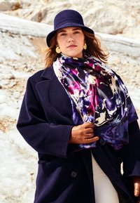 Marineblauer Wollmantel mit Schalkragen, lebendiger Seidenschal mit Blumenmuster in lila Tönen und passendem Filzhut. Das Modell trägt hellbraune Handschuhe.